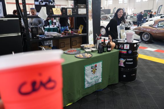 A woman stands behind a table with drinks and snacks at a market, holding a pink cup.