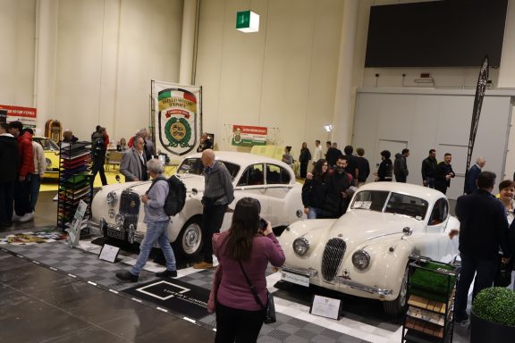 Crowd admiring classic cars at an exhibition, including two vintage white vehicles.