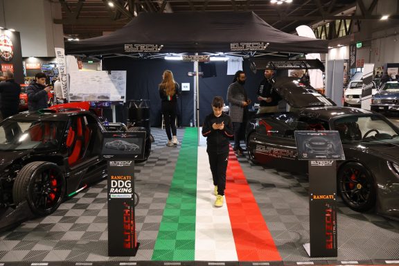 Due persone in un evento automobilistico, con auto sportive e un pavimento tricolore.