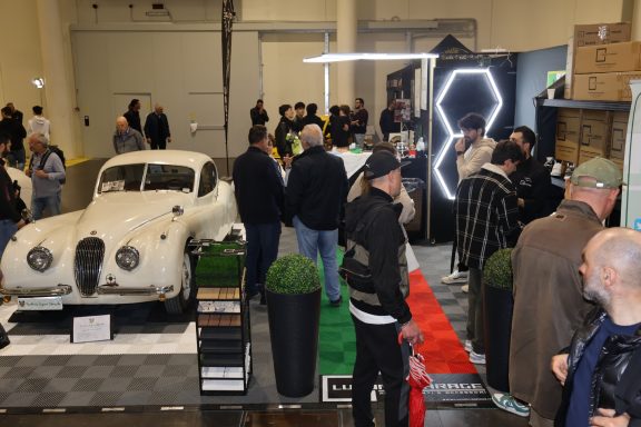 Classic white car on display at an event, surrounded by attendees and greenery.