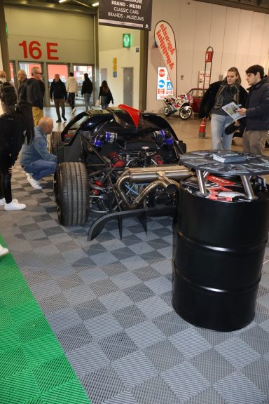 Veicolo da corsa smontato esposto su un pavimento a scacchi, con visitatori intorno.
