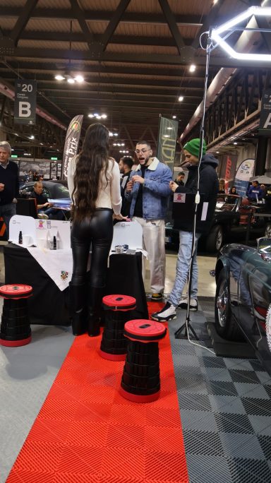 Person with long hair in a white top stands near a booth at a car show. Red carpet and stools visible.