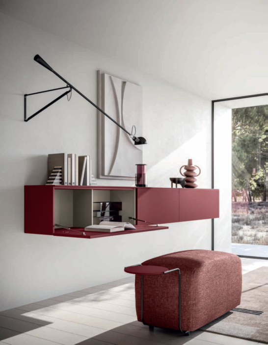 COMPOSIZIONE GIORNO HNG046 Arredamento moderno con parete rossa, lampada, libri e un pouf tessile.