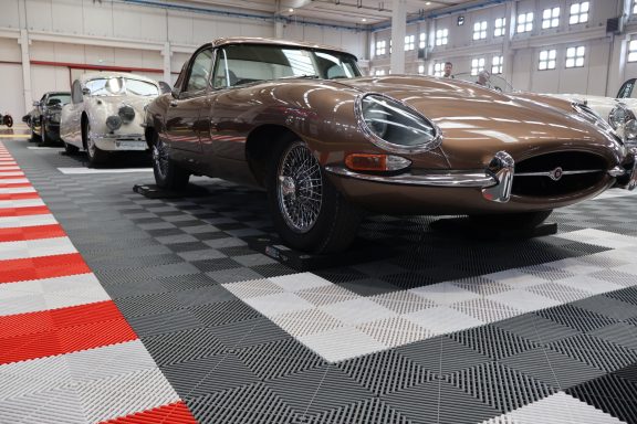 Auto d'epoca marrone, modello E-Type, in esposizione su pavimento a scacchi.