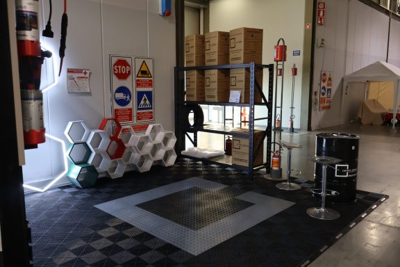 Area in a warehouse with safety signs, stacked boxes, and a shelving unit.