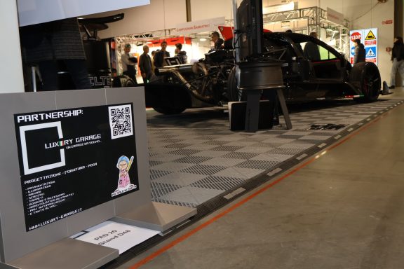 Auto da corsa esposto con un cartello informativo e codice QR in un ambiente di fiera.