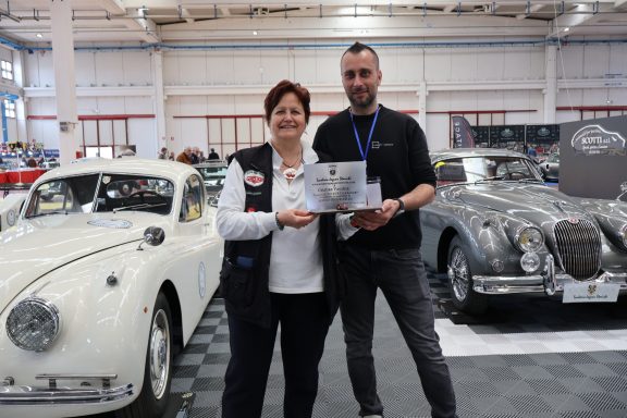 Due persone sorridenti tengono un certificato accanto a due auto d'epoca.