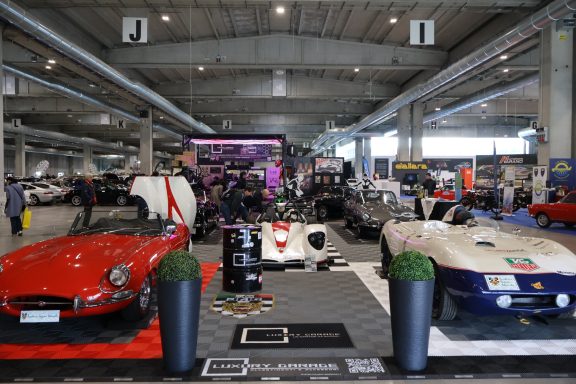 Tre auto sportive d'epoca in mostra, con uno sfondo colorato e un ambiente fieristico.