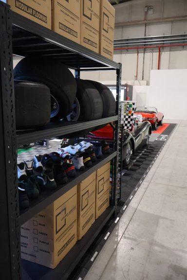 Shelves with boxes and tires beside a red sports car in an industrial setting.