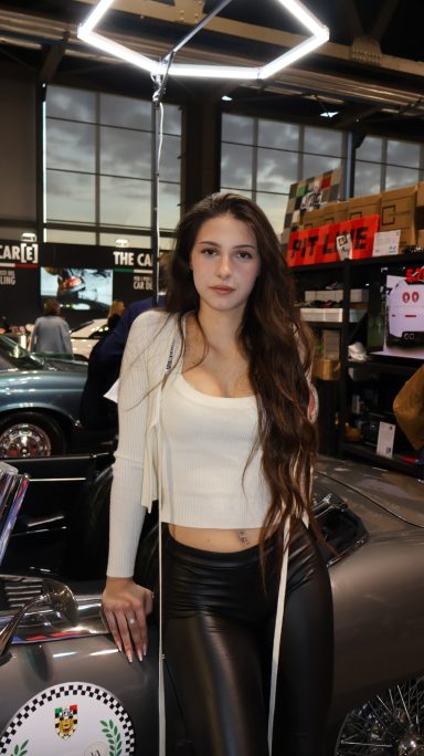 Young woman with long hair wearing a white top and black leather pants, standing near a car.