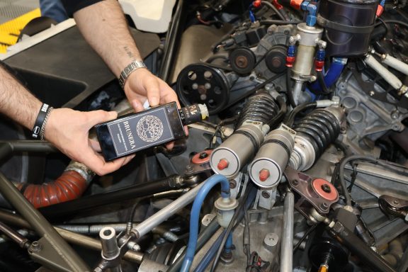 Man holds a bottle over a complex engine assembly with various mechanical components.