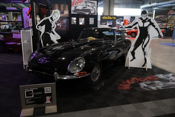 Auto d'epoca nero con sagome bianche di personaggi su sfondo di esposizione.