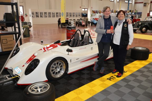 Due due persone sorridenti accanto a una vettura da corsa in un garage.