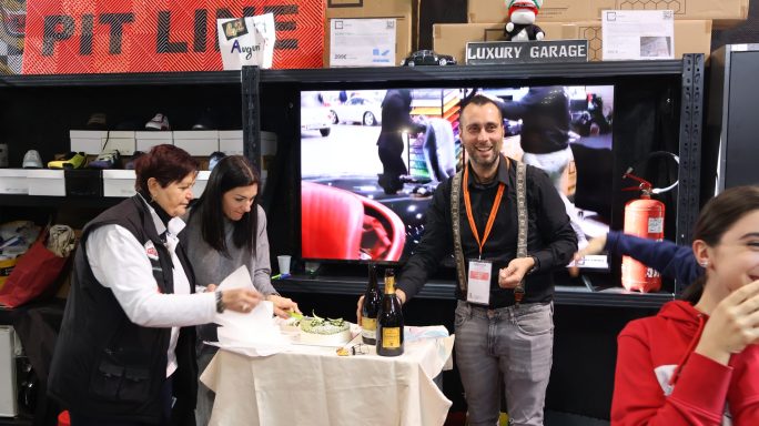 Persone sorridenti interagiscono in un ambiente festivo con cibo e un video sullo sfondo.