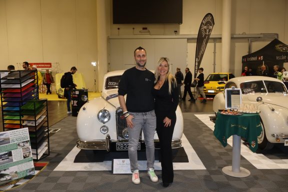 A man and a woman pose together in front of classic cars at an event.